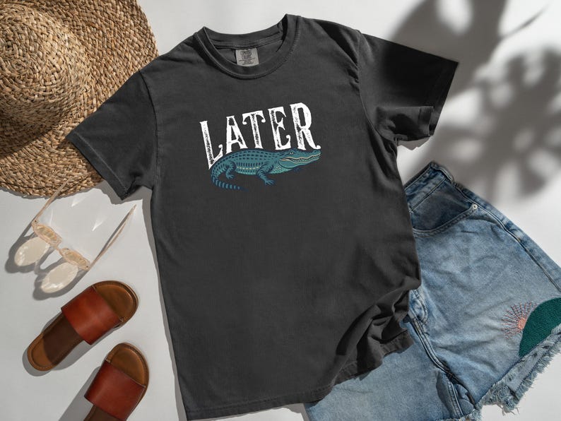 May include: Dark gray t-shirt with the word "LATER" in white, featuring a blue and green alligator illustration. The shirt is paired with denim shorts, sandals, and a straw hat.