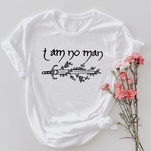 Puede incluir: Camiseta blanca con la frase "i am no man" sobre un dise&ntilde;o de espada y hojas en negro. Un ramo de flores rosas est&aacute; colocado a la derecha de la camiseta.
