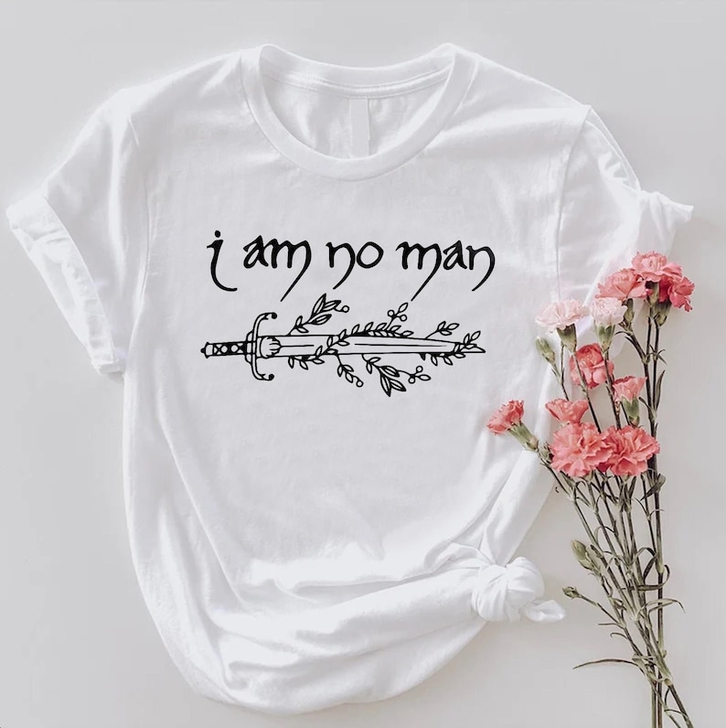 Puede incluir: Camiseta blanca con la frase "i am no man" sobre un dise&ntilde;o de espada y hojas en negro. Un ramo de flores rosas est&aacute; colocado a la derecha de la camiseta.