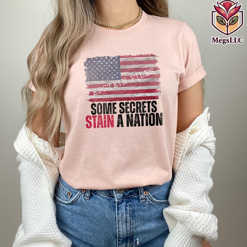 Pode incluir: Camiseta rosa claro com um design desgastado da bandeira americana. A camiseta apresenta o texto "EPSTEIN FILES" acima da bandeira e a frase "SOME SECRETS STAIN A NATION" em preto e vermelho.