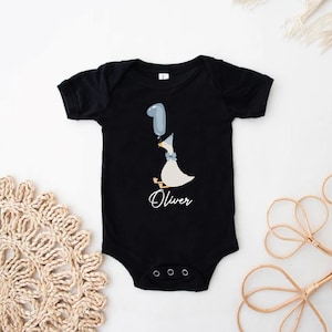 Puede incluir: Body negro con un pato blanco que sostiene un globo azul que dice "1" y el nombre "Oliver" impreso debajo.