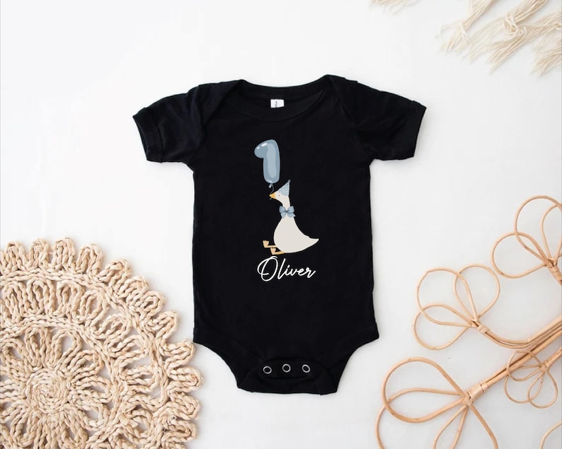 Puede incluir: Body negro con un pato blanco que sostiene un globo azul que dice "1" y el nombre "Oliver" impreso debajo.