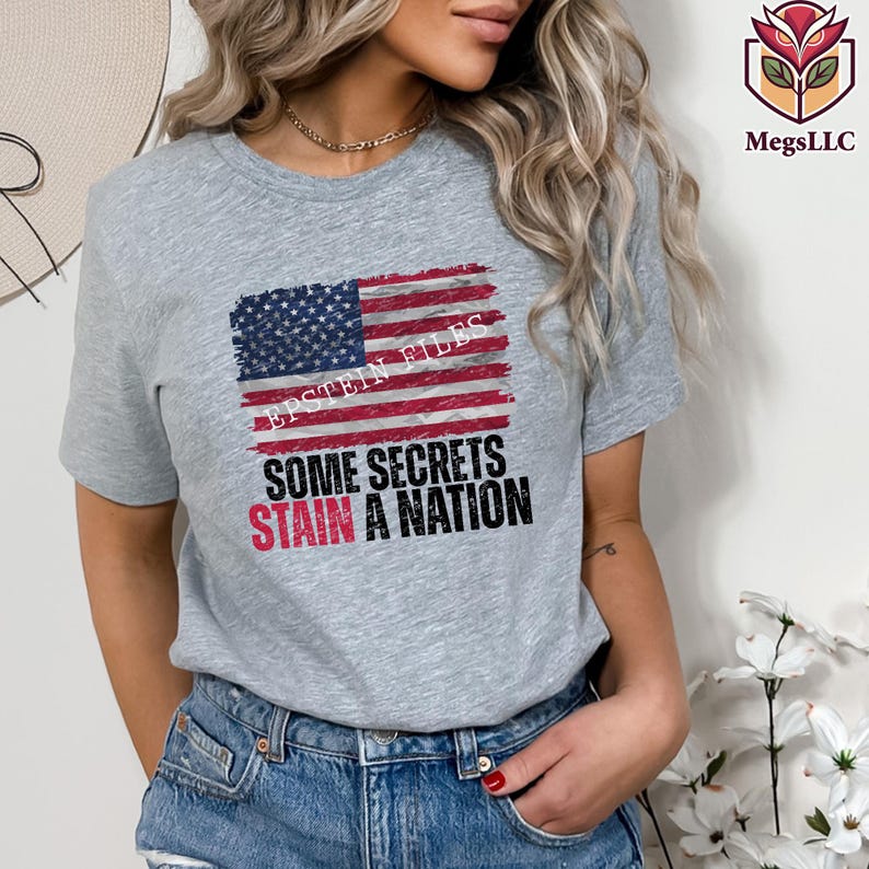 Pode incluir: Uma camiseta cinza com um design desgastado da bandeira americana e o texto "EPSTEIN FILES" acima das palavras "SOME SECRETS STAIN A NATION" em preto e vermelho. Um item de roupa casual.