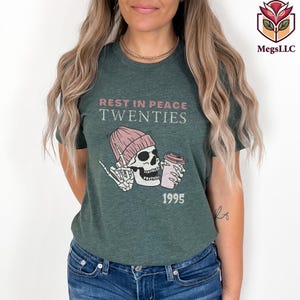 May include: Dark green t-shirt with the text "REST IN PEACE TWENTIES" above a graphic of a skeleton wearing a pink beanie and holding a coffee cup. The year "1995" is printed below the graphic.