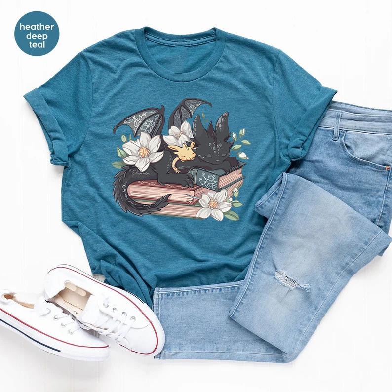 May include: A teal blue t-shirt with a graphic of a black dragon sleeping on a stack of books with a small yellow dragon curled up next to it. The dragon is surrounded by white flowers.