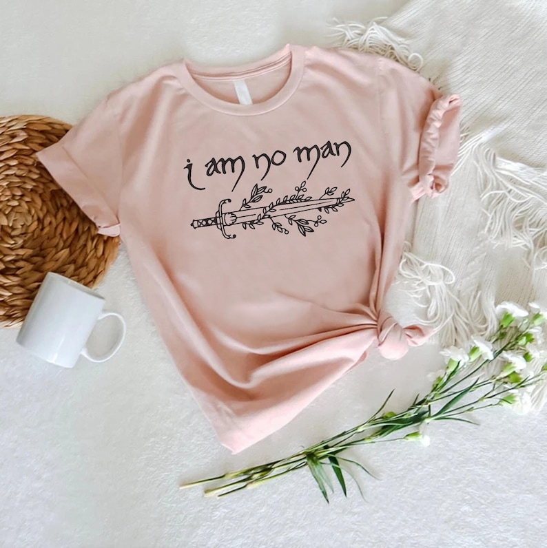 Puede incluir: Camiseta rosa claro con la frase "i am no man" sobre un dise&ntilde;o de espada y hojas. La camiseta est&aacute; anudada en el dobladillo. Una taza blanca, una cesta de mimbre y flores est&aacute;n en el fondo.