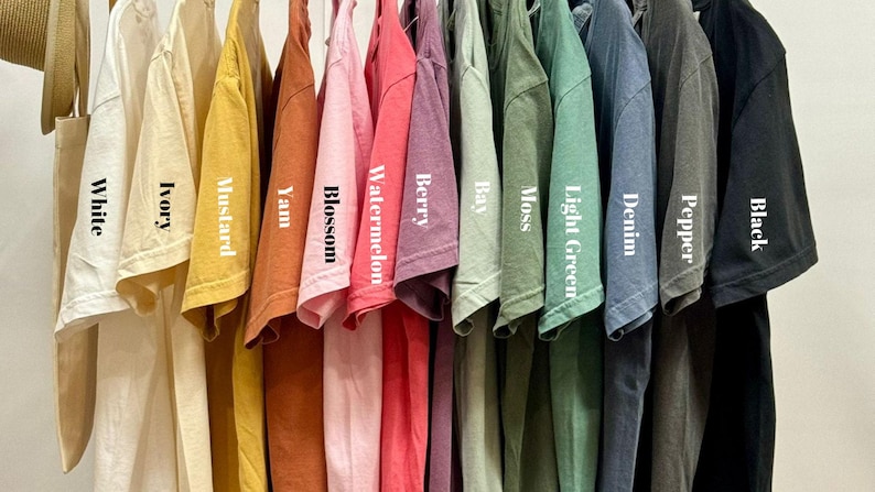 May include: A variety of short-sleeved t-shirts in various colors, including white, ivory, mustard, yam, blossom, watermelon, berry, bay, moss, light green, denim, pepper, and black. Each shirt is labeled with its color name.