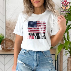 Pode incluir: Camiseta branca com um design desgastado da bandeira americana e o texto "SOME SECRETS STAIN A NATION" em preto e vermelho. A camiseta tem mangas curtas e gola redonda.
