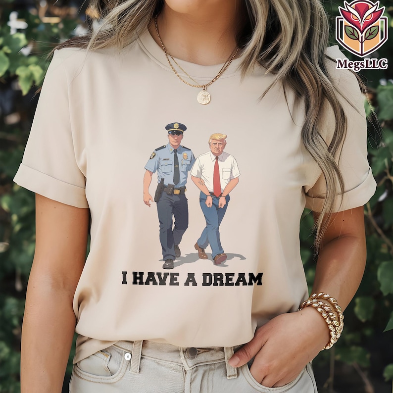May include: Beige t-shirt featuring a cartoon illustration of a police officer and a man in a white shirt and red tie walking together. The text "I HAVE A DREAM" is printed below the image. The shirt has short sleeves.