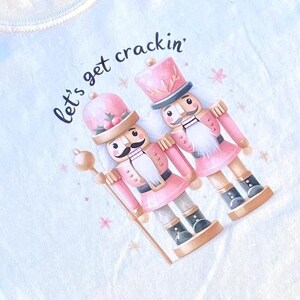 May include: A light blue t-shirt featuring a pink and white illustration of two nutcrackers. The text "Let's get crackin'" is written above the nutcrackers.