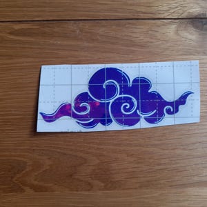 May include: A purple and blue iridescent vinyl decal of a stylised cloud design. The decal is on a white sheet with a grid pattern.