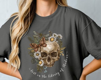 May include: A dark grey t-shirt featuring a floral skull design and the text "Lost in the library of shadows".