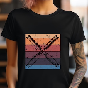 Op de afbeelding: Zwart T-shirt met een afbeelding van twee gekruiste beitels tegen een achtergrond van horizontale strepen in oranje, rood, paars en blauw. Het woord "BLACK" staat onderaan.