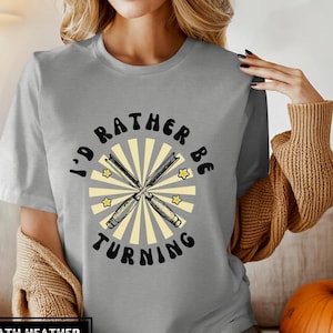 May include: Gray t-shirt with the text "I'd rather be turning" in black lettering. A pair of crossed woodturning chisels are in the center of the design, surrounded by a yellow sunburst with stars.