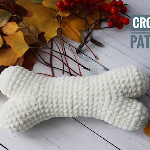 Op de afbeelding: Witte gehaakte hondenbot speelgoed op een wit houten oppervlak met herfstbladeren en rode bessen. De tekst CROCHET PATTERN staat in de rechterbovenhoek van de afbeelding.