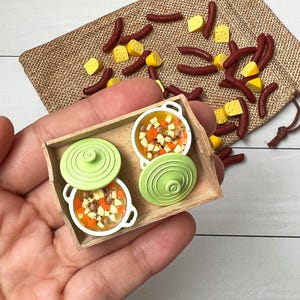 Può includere: Set di cibo in miniatura con due piccole pentole di zuppa con coperchi verdi su un vassoio di legno. Salsicce e cubetti di formaggio in miniatura sparsi, con un sacco di iuta sullo sfondo. Un oggetto artigianale a tema alimentare.
