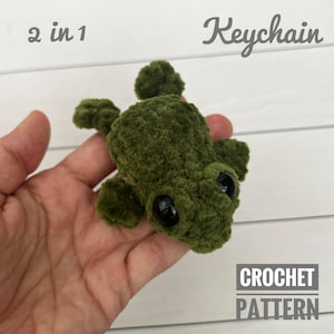 Puede incluir: Un llavero de tortuga verde tejido a ganchillo con grandes ojos negros. El llavero se sostiene en una mano y tiene el texto "2 en 1 Keychain" y "CROCHET PATTERN". La tortuga está hecha de un hilo suave y texturizado.