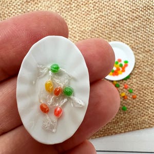 May include: A miniature white oval plate holds wrapped candies in red, green, and yellow. Another small white plate with scattered candies is in the background. The candies are on a textured, tan surface.