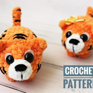 May include: Crochet pattern for two orange and black striped tiger amigurumi plushies with white faces and black eyes. The text "CROCHET PATTERN" is in the bottom right corner of the image.