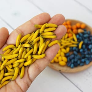 Pode incluir: Uma mão segurando um monte de pequenas bananas em miniatura amarelas. As bananas são feitas de argila polimérica e têm aproximadamente o tamanho de uma unha.