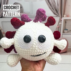May include: White crocheted plush toy with red and purple spikes. The toy has black eyes and a smiling mouth. The toy is being held in a person's hand. CROCHET PATTERN text is visible in the background.
