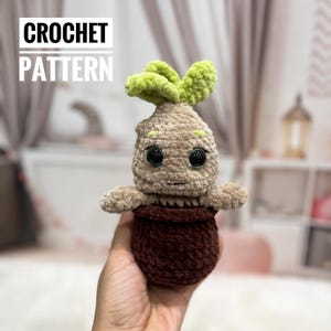 Può includere: Un pupazzo all'uncinetto a forma di Mandragora, con corpo beige, foglie verdi e grandi occhi neri. È seduto in un vaso marrone. Il testo "CROCHET PATTERN" è visualizzato in alto.