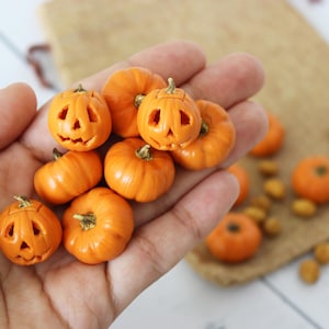 Op de afbeelding: Een hand die een verzameling kleine oranje pompoenen vasthoudt met gesneden jack-o'-lantern gezichten. De pompoenen zijn gemaakt van klei en zijn perfect voor Halloween-decoraties.