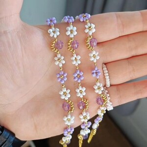 Könnte beinhalten: Drei Armbänder im Rapunzel-Stil mit weißen und lila Blumenperlen, goldenen Akzenten und lila Kristallperlen. Die Armbänder werden in einer Hand gehalten. Der Text auf dem Bild lautet "Rapunzel themed bracelets."