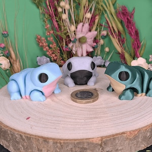 Puede incluir: Tres pequeñas figuritas de rana, una azul, una gris y una verde, están dispuestas sobre una superficie de madera. Una moneda está delante de las ranas.