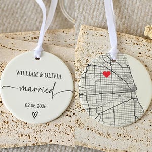 May include: Two round white ceramic ornaments with white ribbon hangers. One side features the text WILLIAM & OLIVIA, married, and the date 02.06.2026 with a small heart. The other side displays a black street map with a red heart icon.