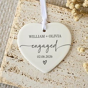 May include: A white heart-shaped ceramic ornament featuring the printed text WILLIAM + OLIVIA, the word engaged in a cursive script, and the date 02.06.2026 with a small heart icon below. A thin white ribbon is attached through a hole at the top.