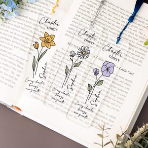 Op de afbeelding: Transparante acryl bladwijzers met bloemmotieven en kwastjes. Elke bladwijzer heeft een andere bloemillustratie: narcis, madeliefje en viooltje. Tekst bevat "Chapter Thirty" en de naam "Sophia".