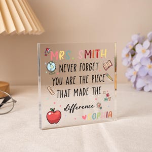 May include: A clear acrylic block with colorful text and illustrations. The text reads "MRS. SMITH NEVER FORGET YOU ARE THE PIECE THAT MADE THE difference SOPHIA." Includes a red apple, globe, and pencil.