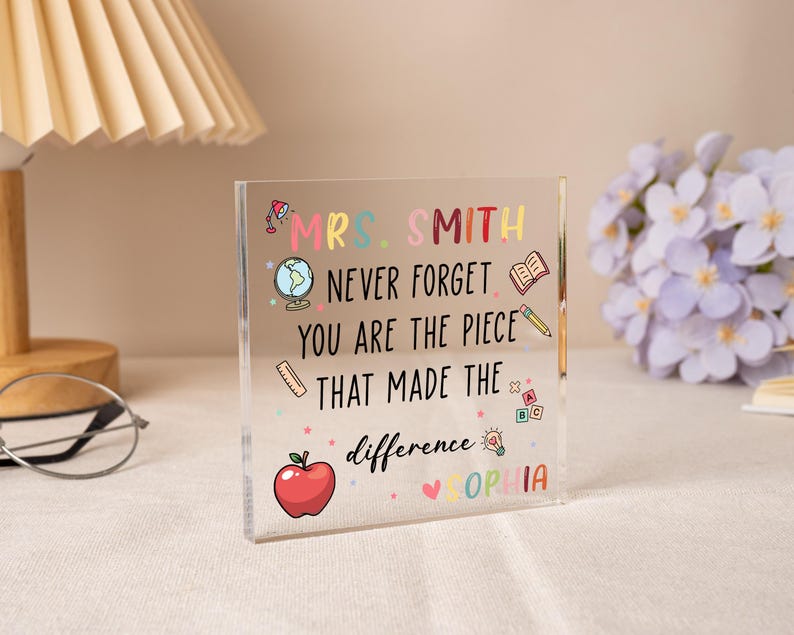 May include: A clear acrylic block with colorful text and illustrations. The text reads "MRS. SMITH NEVER FORGET YOU ARE THE PIECE THAT MADE THE difference SOPHIA." Includes a red apple, globe, and pencil.