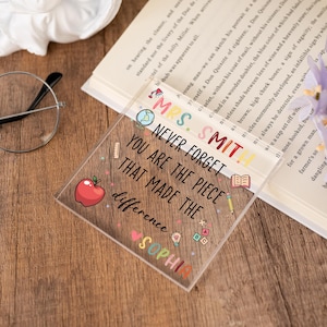 May include: A clear acrylic teacher appreciation gift with the text "Mrs. Smith, Never Forget You Are The Piece That Made The Difference, Sophia." The square-shaped gift has colorful illustrations of an apple, pencil, and globe.