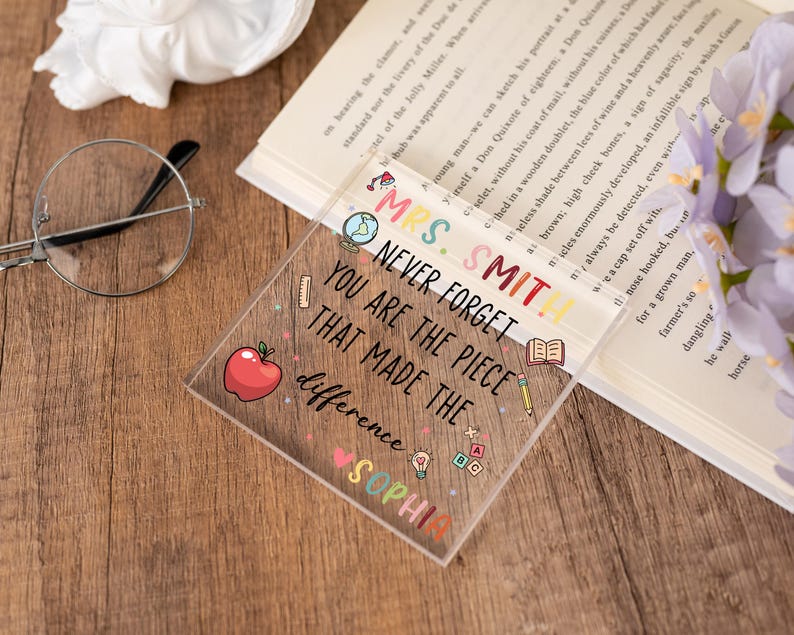 May include: A clear acrylic teacher appreciation gift with the text "Mrs. Smith, Never Forget You Are The Piece That Made The Difference, Sophia." The square-shaped gift has colorful illustrations of an apple, pencil, and globe.