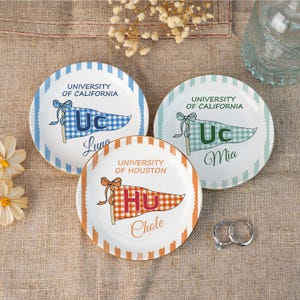 May include: Three decorative ceramic plates with university-themed designs. Two plates feature "University of California" with blue and green accents, and the third plate displays "University of Houston" with orange details. Each plate has a gold rim.