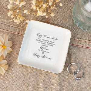 May include: A white square ceramic trinket dish with gold trim. The dish has the text "Enjoy the next chapter" and "Happy Retirement" in black script. Two silver rings are next to the dish.