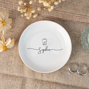 Op de afbeelding: Een witte keramische ringenschotel met een gouden rand. De schotel heeft een zwarte omtrek van een tand en de naam "Sophia" in cursief.