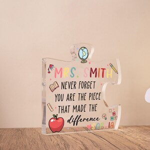 May include: Clear acrylic puzzle piece with the text "MRS. SMITH NEVER FORGET YOU ARE THE PIECE THAT MADE THE difference". The design includes a red apple, a globe, a pencil, and other school-themed illustrations.