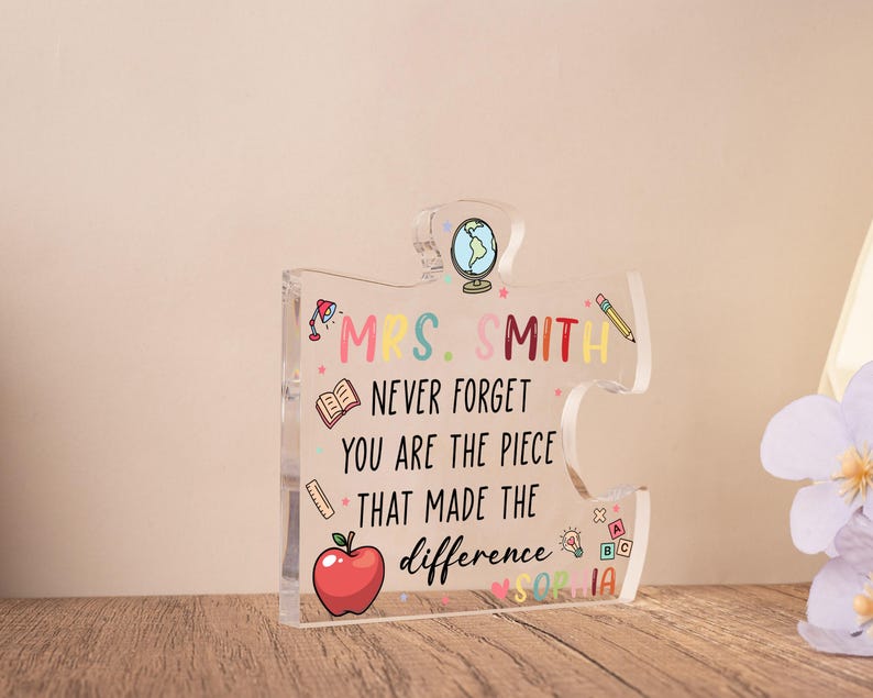 May include: Clear acrylic puzzle piece with the text "MRS. SMITH NEVER FORGET YOU ARE THE PIECE THAT MADE THE difference". The design includes a red apple, a globe, a pencil, and other school-themed illustrations.