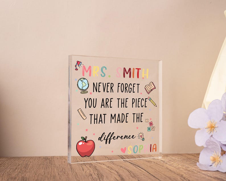 May include: A clear acrylic block with colorful text and illustrations. The text reads "MRS. SMITH NEVER FORGET YOU ARE THE PIECE THAT MADE THE difference SOPHIA." Includes a red apple, globe, and pencil.