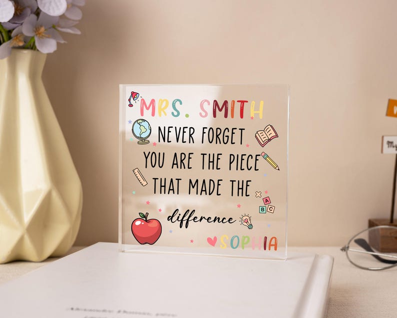 May include: A clear acrylic block with colorful text and illustrations. The text reads "MRS. SMITH NEVER FORGET YOU ARE THE PIECE THAT MADE THE difference SOPHIA". Includes a globe, apple, book, pencil, and other school-themed icons.
