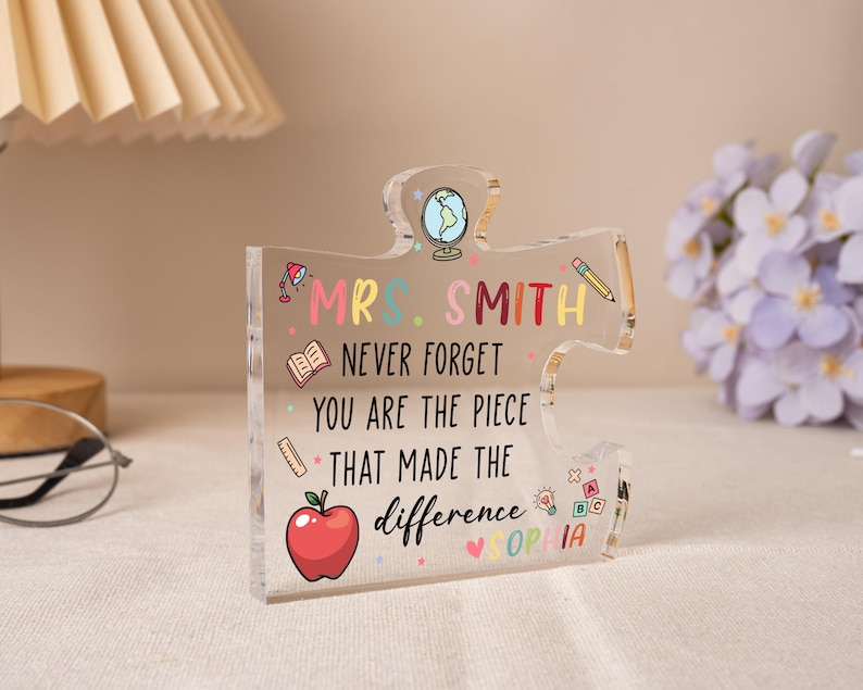 May include: A clear acrylic puzzle piece with the text "MRS. SMITH NEVER FORGET YOU ARE THE PIECE THAT MADE THE difference SOPHIA" in colorful letters. The design includes a globe, apple, and school-themed icons.
