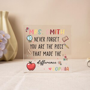 May include: A clear acrylic block with colorful text and illustrations. The text reads "MRS. SMITH NEVER FORGET YOU ARE THE PIECE THAT MADE THE difference SOPHIA." Includes an apple, globe, book, and pencil.