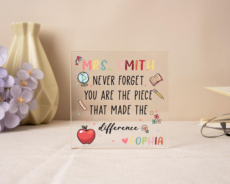 May include: A clear acrylic block with colorful text and illustrations. The text reads "MRS. SMITH NEVER FORGET YOU ARE THE PIECE THAT MADE THE difference SOPHIA." Includes an apple, globe, book, and pencil.