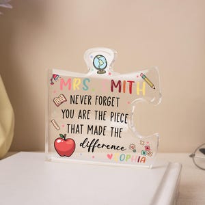 May include: A clear acrylic puzzle piece with the text "MRS. SMITH NEVER FORGET YOU ARE THE PIECE THAT MADE THE DIFFERENCE SOPHIA". The puzzle piece has colorful illustrations of a globe, pencil, apple, and other school-related icons.