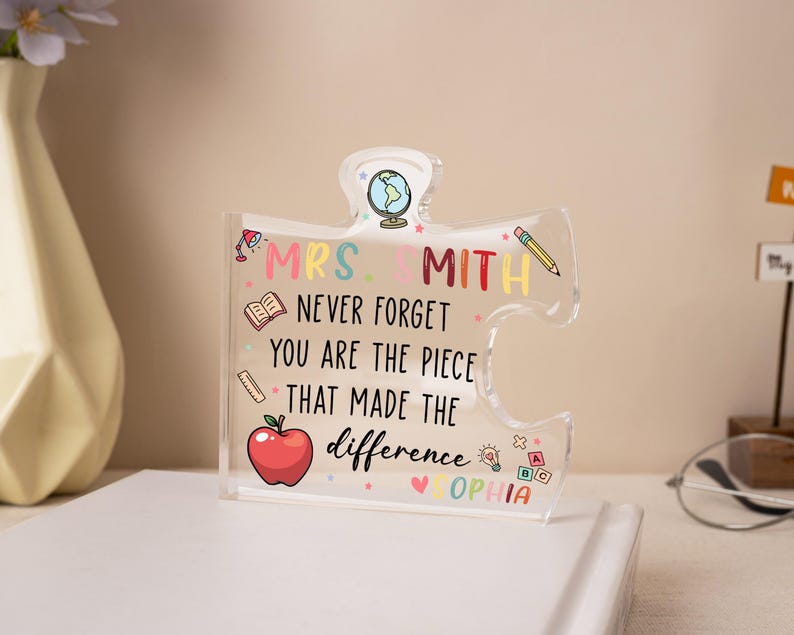 May include: A clear acrylic puzzle piece with the text "MRS. SMITH NEVER FORGET YOU ARE THE PIECE THAT MADE THE DIFFERENCE SOPHIA". The puzzle piece has colorful illustrations of a globe, pencil, apple, and other school-related icons.