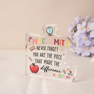 May include: A clear acrylic puzzle piece with the text "MRS. SMITH NEVER FORGET YOU ARE THE PIECE THAT MADE THE difference OPHIA". The design includes a globe, apple, pencil, and book illustrations. A thoughtful teacher gift.