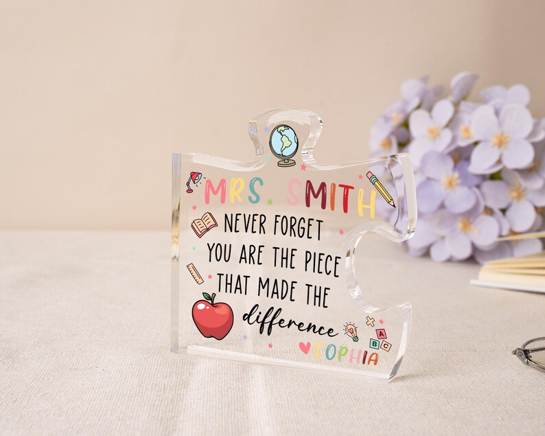 May include: A clear acrylic puzzle piece with the text "MRS. SMITH NEVER FORGET YOU ARE THE PIECE THAT MADE THE difference OPHIA". The design includes a globe, apple, pencil, and book illustrations. A thoughtful teacher gift.